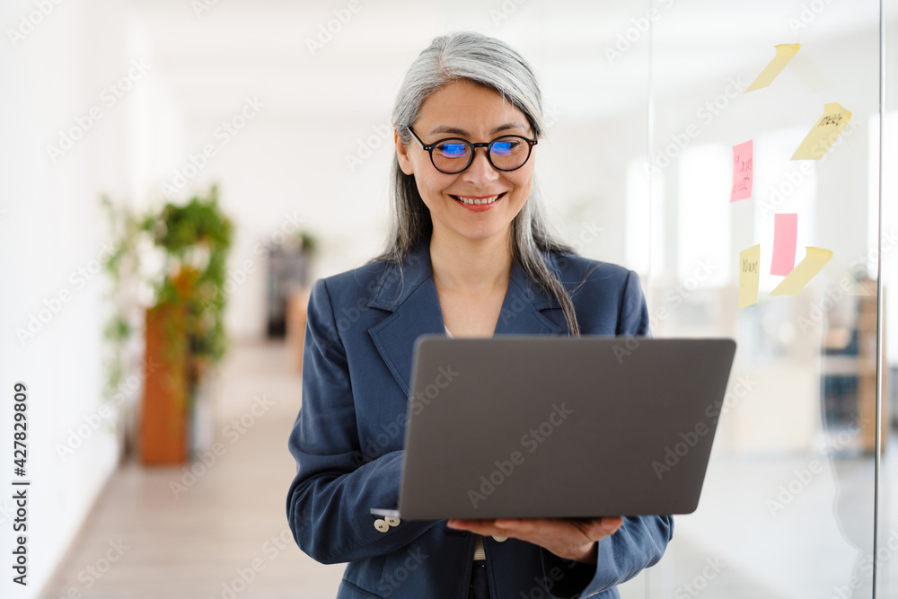 The smiling Asian woman in glasses holding a laptop in her hands while standing in a bright office The smiling Asian woman in glasses holding a laptop in her hands while standing in a bright office