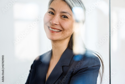 The portrait of happy Asian woman standing behind glass and looking to the camera in the office