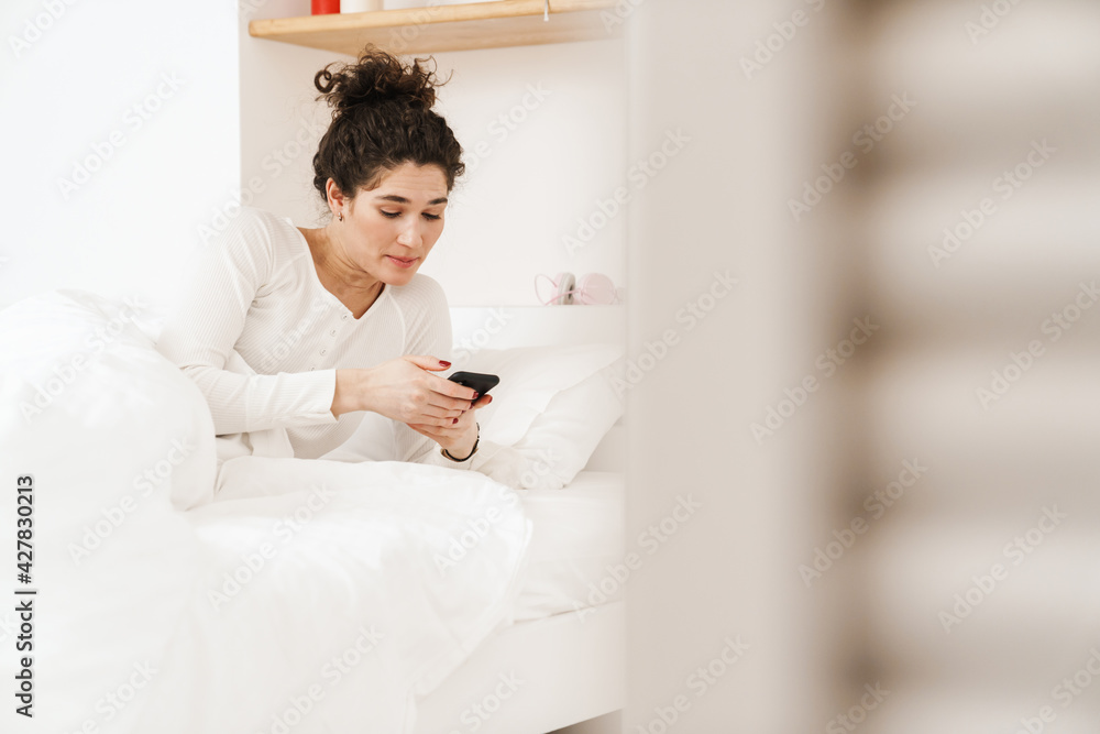 The surprised woman looking at the phone while lying on the bed in the room The surprised woman looking at the phone while lying on the bed in the room