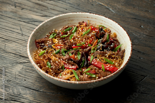 Delicious Asian street food - funchose noodles with beef, cilantro, vegetables and omelet in a white bowl on a wooden background. Cellophane noodles wok