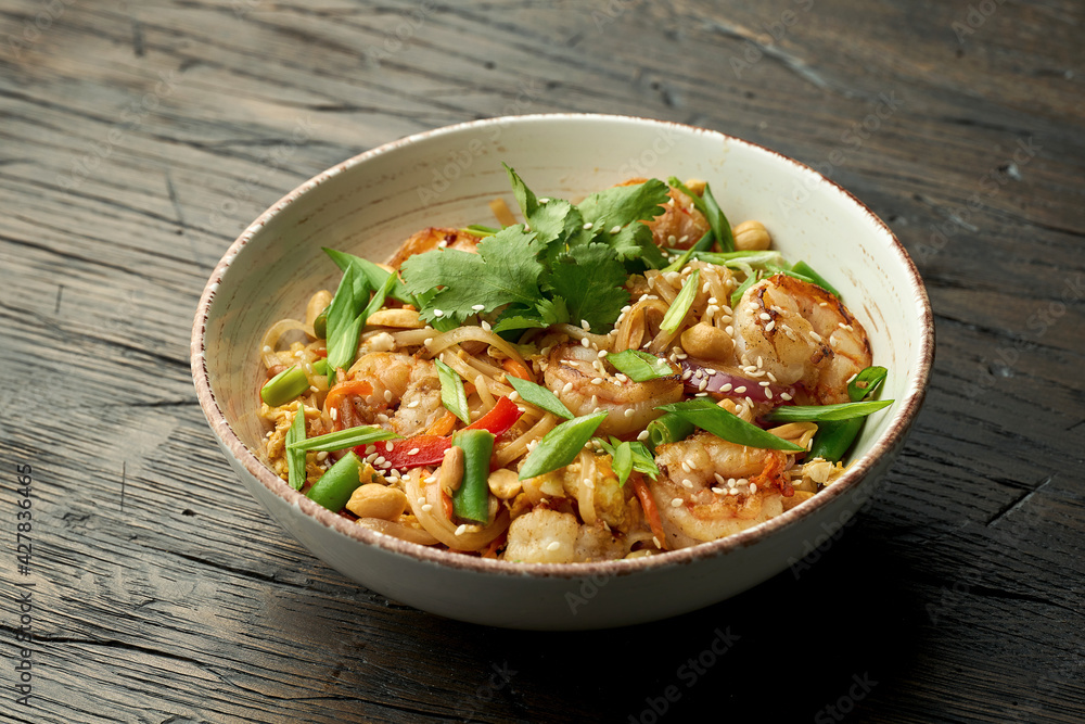 Delicious asian street food - pad thai noodles with shrimp, cilantro, vegetables and scrambled eggs in a white bowl on a wooden background.