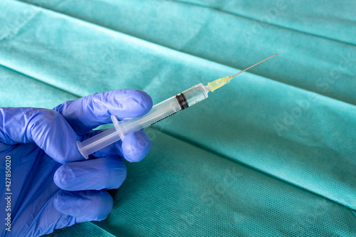 Health Care worker with blue surgical gloves holding a syringe in front of a green surgical fabric. Surgeon hand with a vaccine. A syringe with the vaccine. 