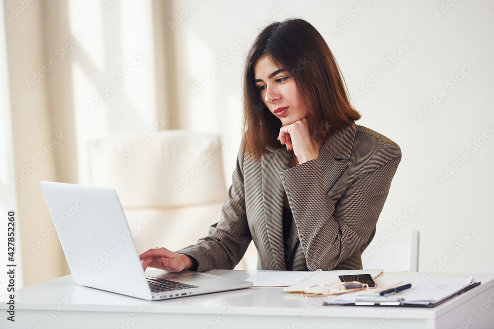Works in the office by laptop. Young woman in formal clothes is indoors. Conception of style Works in the office by laptop. Young woman in formal clothes is indoors. Conception of style