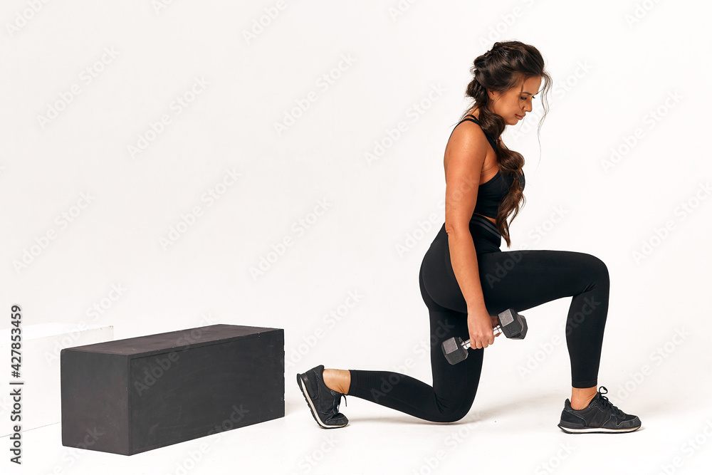 Fototapeta premium Athletic young woman in a black suit lunges forward with dumbbells on a white background. crossfit. fitness squats healthy lifestyle