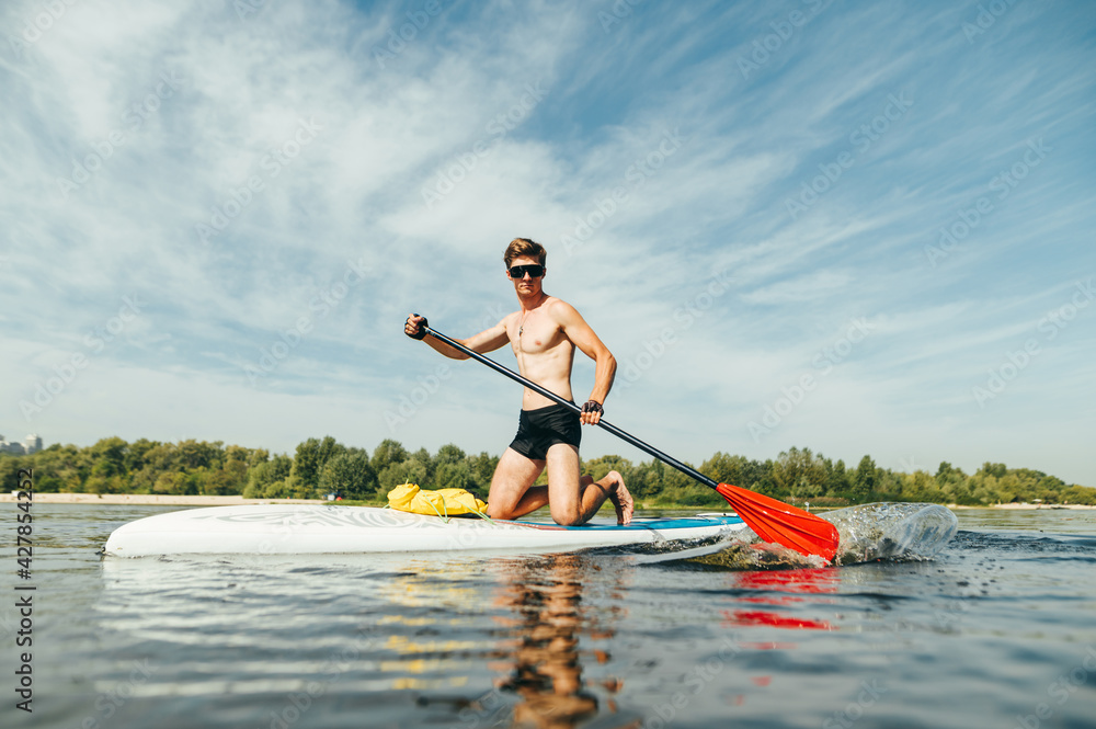 Muscular athletic man in sunglasses actively rowing with an oar sitting on a sup board, swims on a pond. Summer fun on the water.