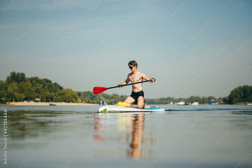 Muscular athlete trains in swimming on the river on a sup board, actively rowing with a paddle with a serious face