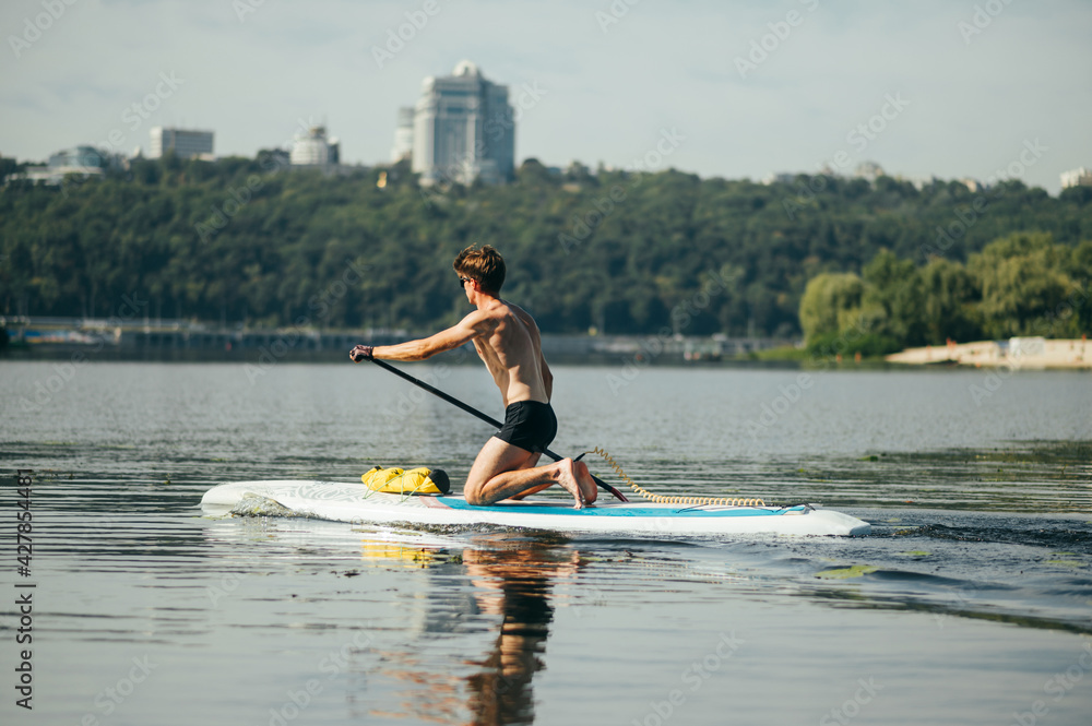 Athletic young man surfing on a sup board on the river in the summer on a weekend, actively rowing, rear view.