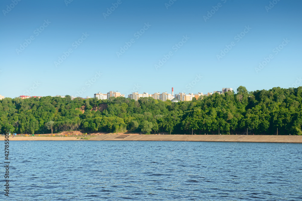 Summer landscape with a river bank with a concrete bank, green forest and high-rise buildings