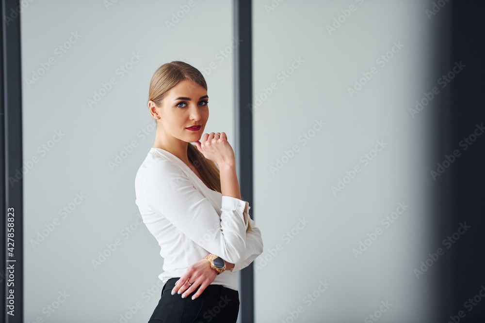Standing against grey background. Young adult woman in formal clothes is indoors in the office Standing against grey background. Young adult woman in formal clothes is indoors in the office