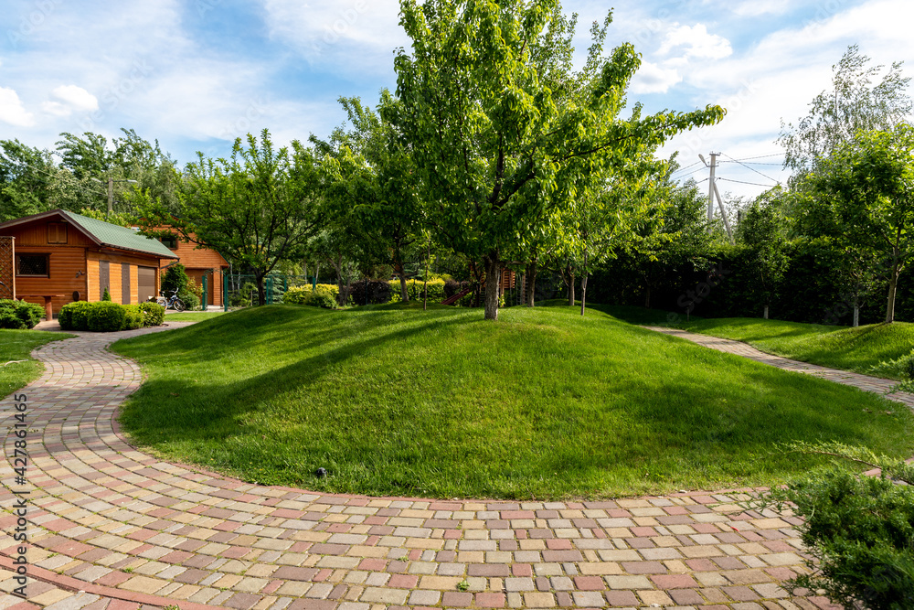 Scenic view yard garden trees and paved stone path road for walk against beautiful blue sky. Landscape design green lawn turf hills and plants irrigated with smart autonomous sprayers at bright day