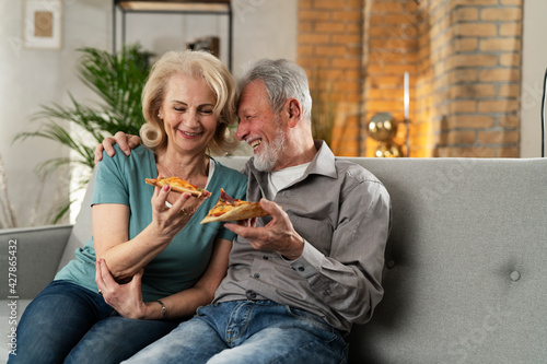 Cheerful husband and wife sitting on sofa at home. Happy senior woman and man eating pizza while watching a movie...