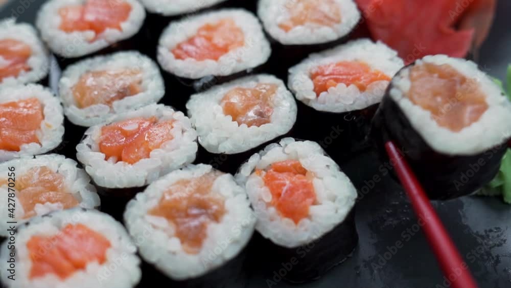 Close-up of Eating Sushi, Roll with a Black Dish. Red Sticks Take Sushi, a Roll of Salmon and Rice. The concept of Healthy Seafood Food. Close-up of Eating Sushi, Roll with a Black Dish. Red Sticks Take Sushi, a Roll of Salmon and Rice. The concept of Healthy Seafood Food.