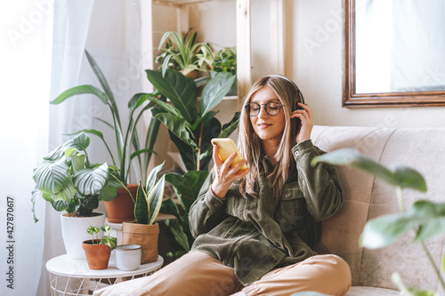 Freelance woman in glasses with mobile phone listening music in headphones and relax at home. Happy girl with closed eye sitting on couch in living room with plants. 
