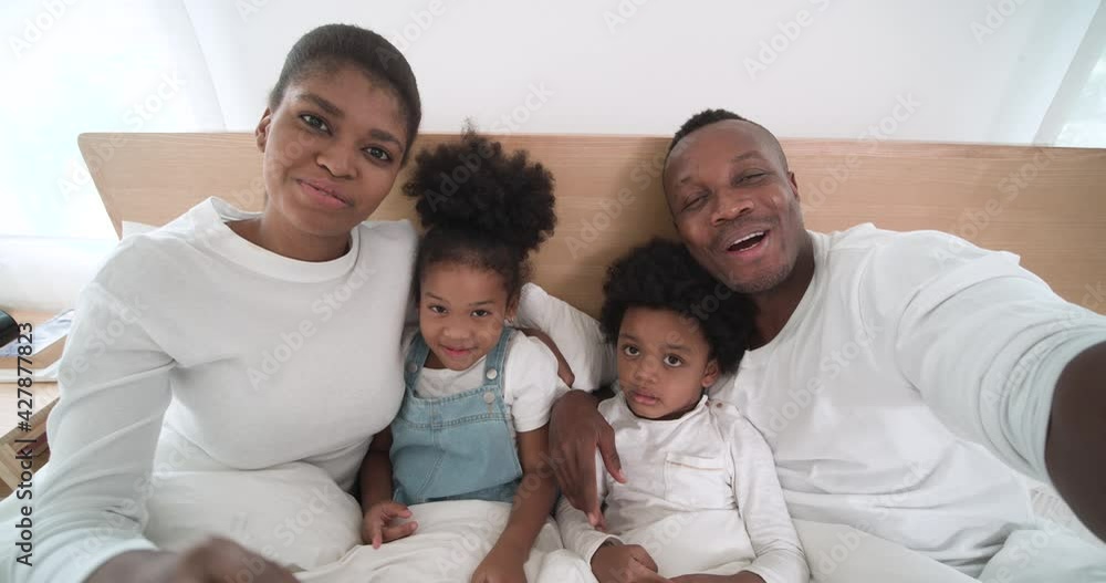 POV shot of Happy African American family lying on bed in bedroom and making video call to talking with their cousin. Young parent using smartphone to record video with their adorable kids at home. POV shot of Happy African American family lying on bed in bedroom and making video call to talking with their cousin. Young parent using smartphone to record video with their adorable kids at home.