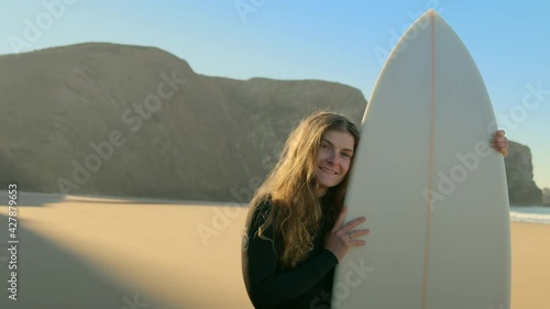 Portrait of beautiful millennial young woman smile to camera. Female surfer stand with surfboard on epic sun filled beach before go into water. Candid and authentic real person doing sports outdoors
