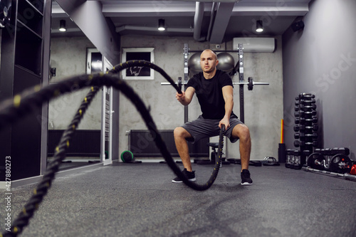 Man training with battle rope in cross fit gym. Strong muscular man working out with battle ropes. Side view photo of fitness man in fashionable sportswear. Strength and motivation