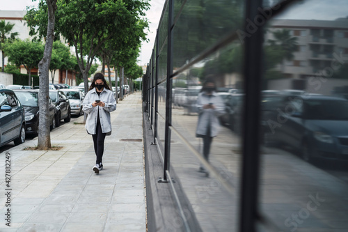 Chica joven con mascarilla negra por covid 19 usando el smartphone en la calle junto a una pared negra