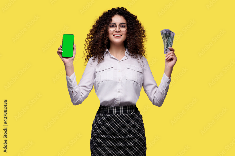 Happy business woman with mobile phone with blank green screen and bunch of dollars over isolated yellow background. Female won money on bet, casino, lottery Happy business woman with mobile phone with blank green screen and bunch of dollars over isolated yellow background. Female won money on bet, casino, lottery
