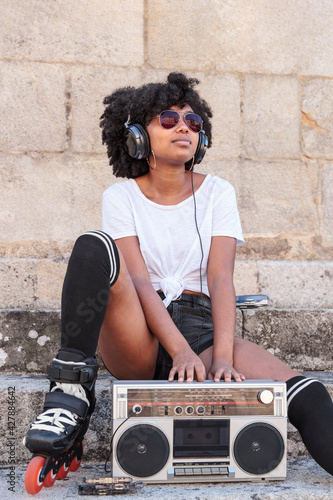 Portrait of a black woman with afro hair, roller skates and sunglasses listening to music with headphones from an old radio cassette player while sitting on a stone stairs outdoors on a hot summer day
