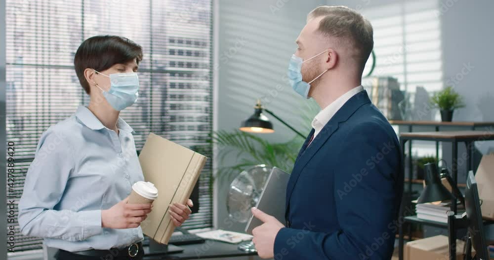 Two managers: man and woman, in protective masks meeting in office and knock their elbows greeting, keeping social distance. Two managers: man and woman, in protective masks meeting in office and knock their elbows greeting, keeping social distance.