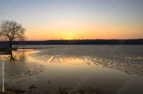 Golden sunset on a lake with tree silhouette and small pier in the foreground. Clear sky and trees in the background. Remains of half-melted ice form fancy pattern on the water.