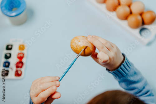 A little child paints an easter egg at the table on a blue background.Little Boy painting Easter Eggs with Paint at Home. Children's Easter creativity. A child decorate Easter eggs.