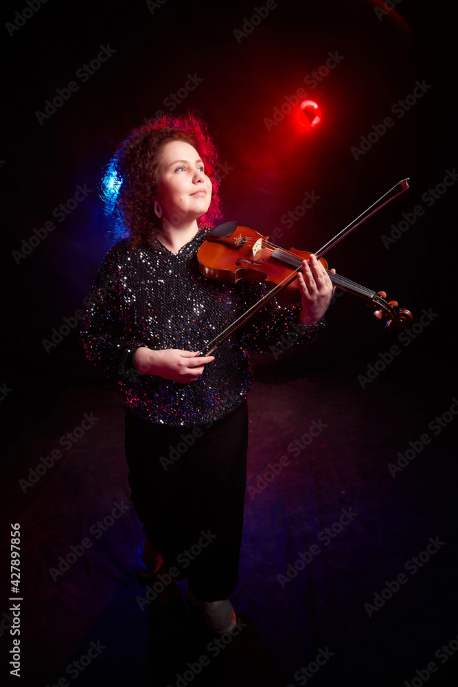 Fototapeta premium A brunette curly girl in black shiny clothes and with violin on a black background. Model, actress and violinist posing in the studio