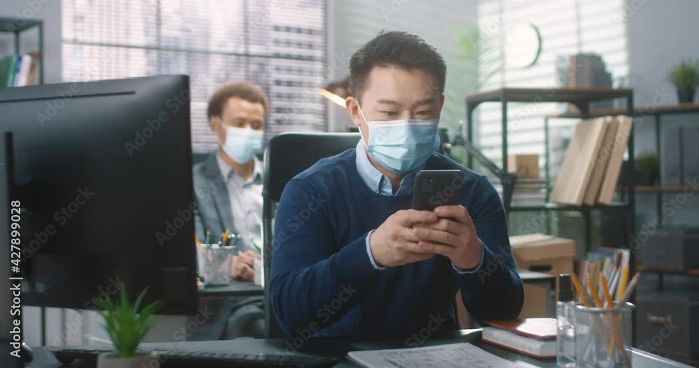 Handsome Asian worker in protective medical mask sitting in office and chatting with friends on social networks using smartphone. Handsome Asian worker in protective medical mask sitting in office and chatting with friends on social networks using smartphone.