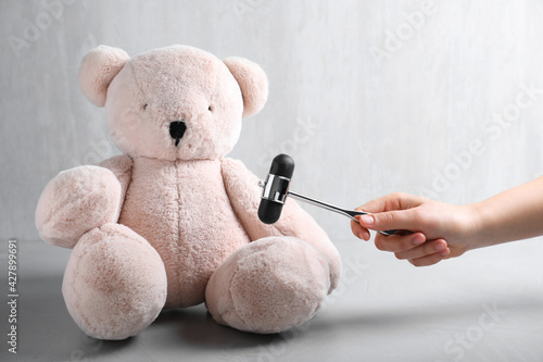 Woman pretending to test teddy bear's reflexes with hammer on grey background, closeup. Nervous system diagnostic photo