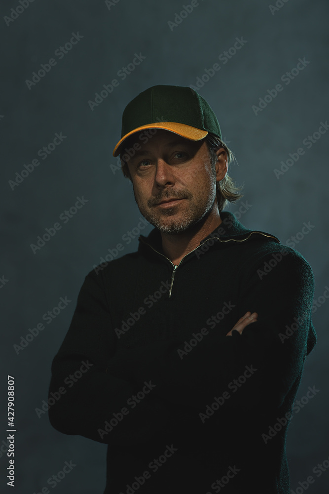 Smiling blond man with a baseball cap, stubble beard and crossed arms in a dark green wool sweater in front of a grey wall. Smiling blond man with a baseball cap, stubble beard and crossed arms in a dark green wool sweater in front of a grey wall.