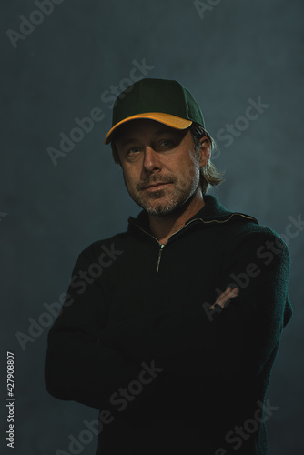 Smiling blond man with a baseball cap, stubble beard and crossed arms in a dark green wool sweater in front of a grey wall. © ysbrandcosijn