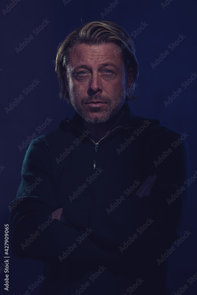 Fototapeta premium Shaded dark portrait of a blond man with stubble beard in a dark green wool sweater with crossed arms in front of a dark blue wall.