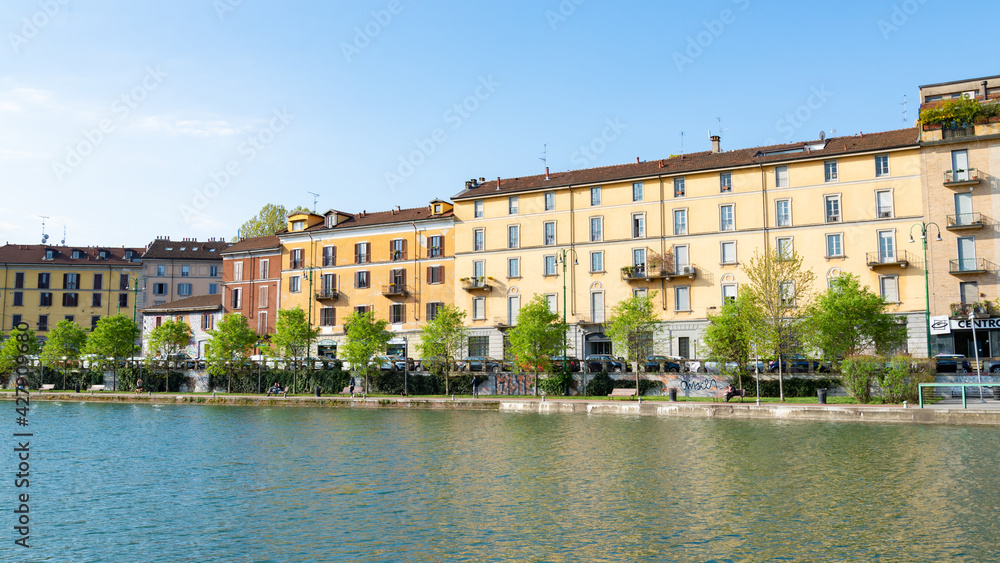 The famous Darsena (meaning: dock) in Milan, Italy, part of the old Navigli neighborhood. Water canal in the foreground, looking like a river. Buildings and blue sky in the background.