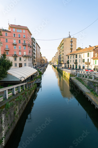 Perspective of the famous old Navigli neighborhood, Milan, Italy. Water canal in the foreground, looking like a river. Buildings and blue sky in the background. © Travelling Jack