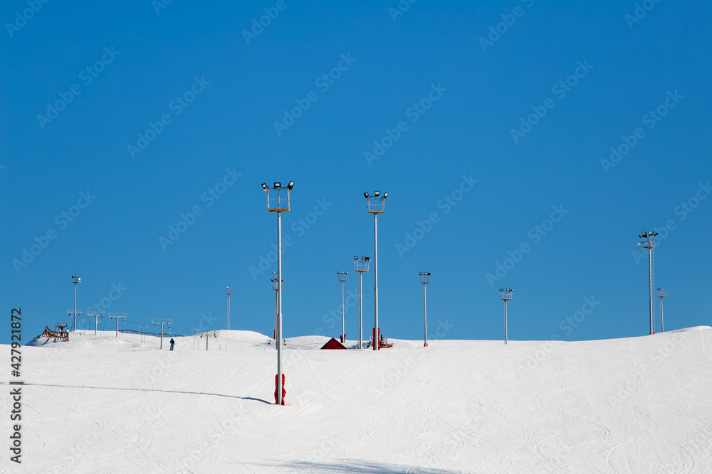 Ski resort, snow slope, trail with artificial lighting towers. Mountain slope for skiing and snowboarding. Against the background of the blue sky. The summit of the rise and ski tours on a sunny day. Ski resort, snow slope, trail with artificial lighting towers. Mountain slope for skiing and snowboarding. Against the background of the blue sky. The summit of the rise and ski tours on a sunny day.