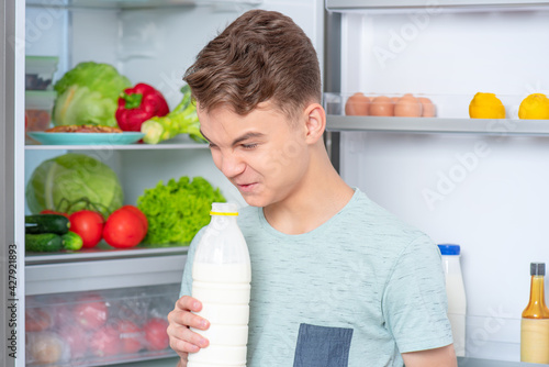 Cute young teen boy holding bottle of milk and drinks while standing near open fridge in kitchen at home. Portrait of pretty child choosing food in refrigerator full of healthy products