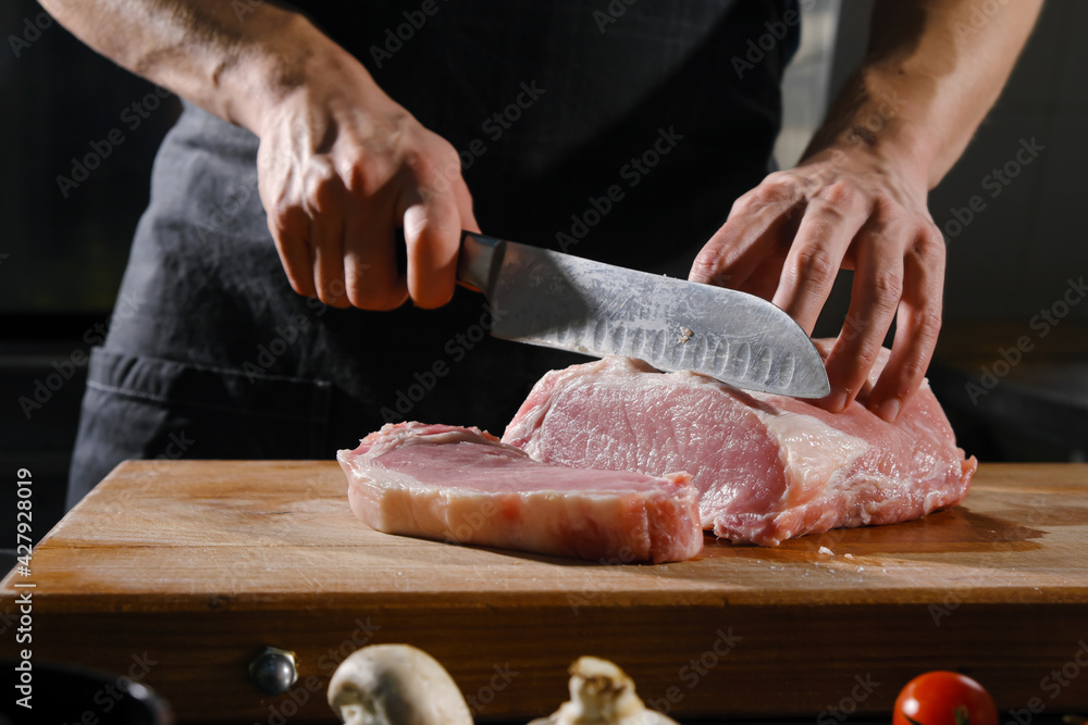 The male chef cuts raw meat with a knife on a wooden board. The restaurant kitchen cooking.
