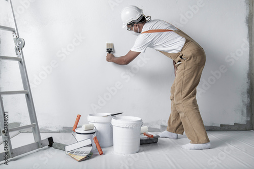 a man paints a white wall with a roller. Repair of the interior. Young male decorator painting a wall in the empty room, concept builder or painter in helmet with paint roller over the empty room