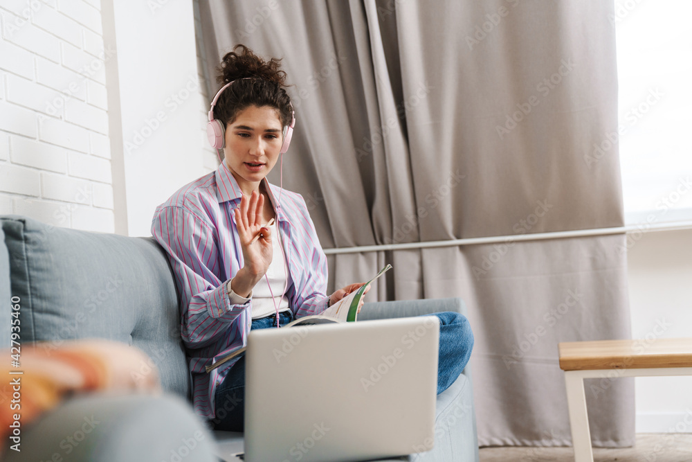 The woman sitting in front of a laptop in headphones and waving to his camera in a room The woman sitting in front of a laptop in headphones and waving to his camera in a room