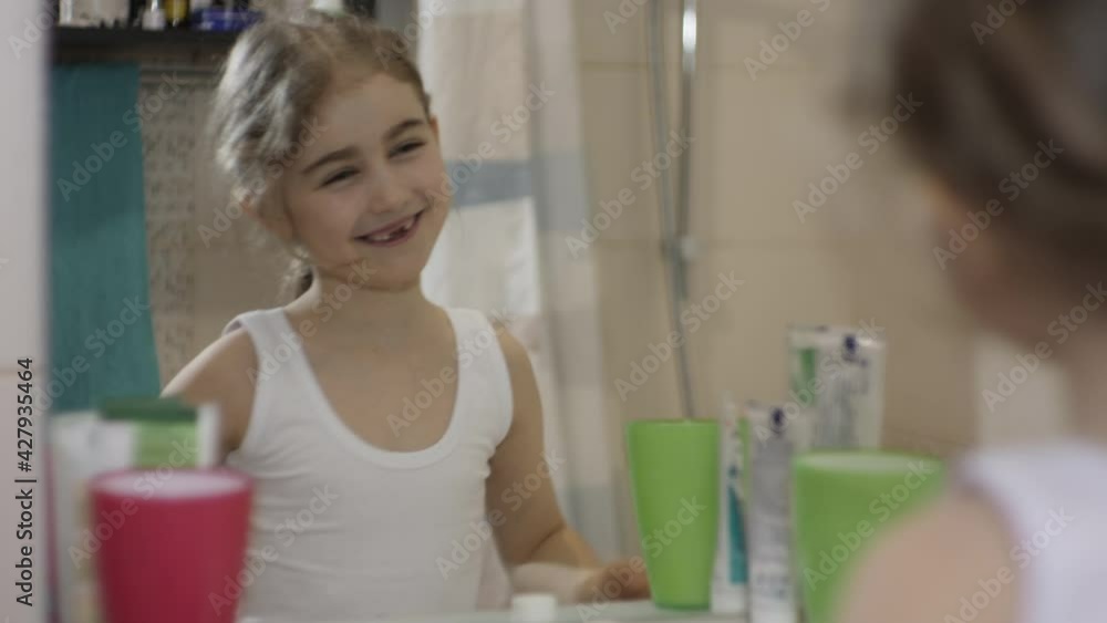 Close-up Portrait Young Little Girl Dancing and Singing in Bathroom Looking at Mirror Reflection. Daily Routine Dental Hygiene. Happy Portrait Caucasian Toothless Child Dancing in Bathroom. Close-up Portrait Young Little Girl Dancing and Singing in Bathroom Looking at Mirror Reflection. Daily Routine Dental Hygiene. Happy Portrait Caucasian Toothless Child Dancing in Bathroom.