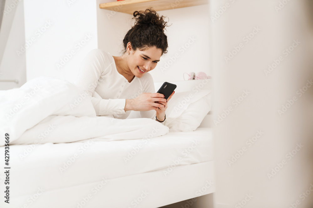 The smiling woman looking at the phone while lying on the bed in the room The smiling woman looking at the phone while lying on the bed in the room