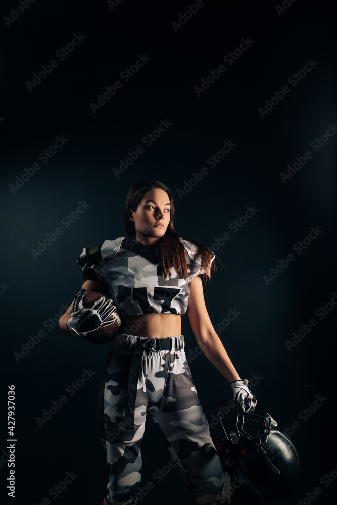 Athletic woman poses in American football uniform with helmet and ball in her hands.