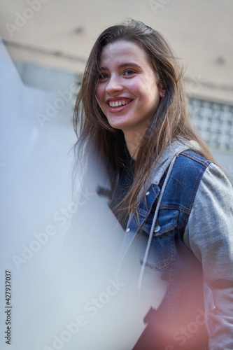 Pretty girl with fair skin and long hair smiling with perfect smile in blue denim jacket and black t-shirt with city in the background. smiling teenager