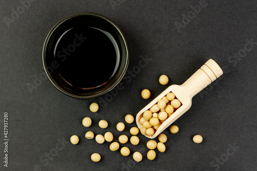 Soy sauce in small black bowl with soybeans on wooden spoon on black background, top view. Soya sauce as a cooking ingredient.