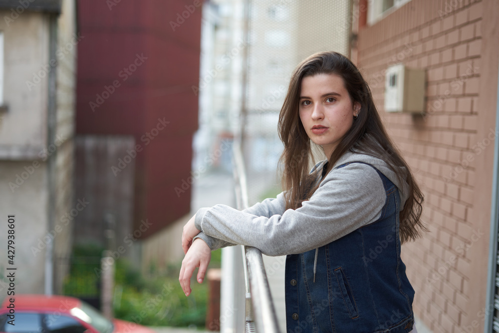 Young girl with long hair and light skin wearing a blue denim jacket leaning on a railing looking at the camera with unfocused urban background. teenager with street look in a city