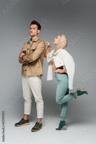 full length of stylish model in pants and jacket pointing at man posing on grey