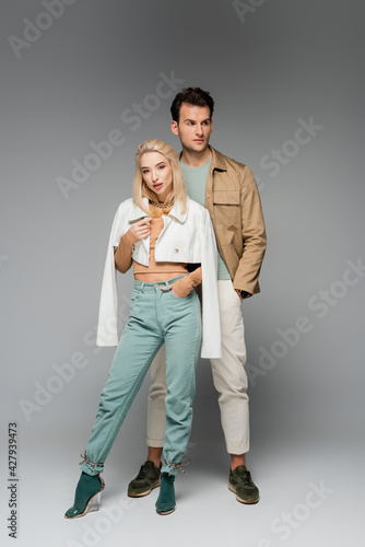 full length of trendy models in pants and jackets posing with hands in pockets on grey