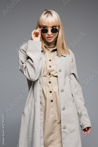 trendy young woman in trench coat adjusting sunglasses isolated on grey