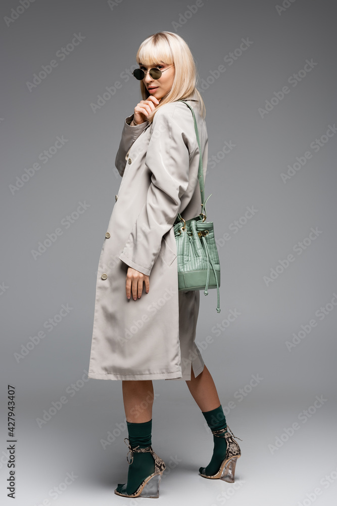 full length of young woman in sunglasses and trench coat with green bag standing on grey full length of young woman in sunglasses and trench coat with green bag standing on grey