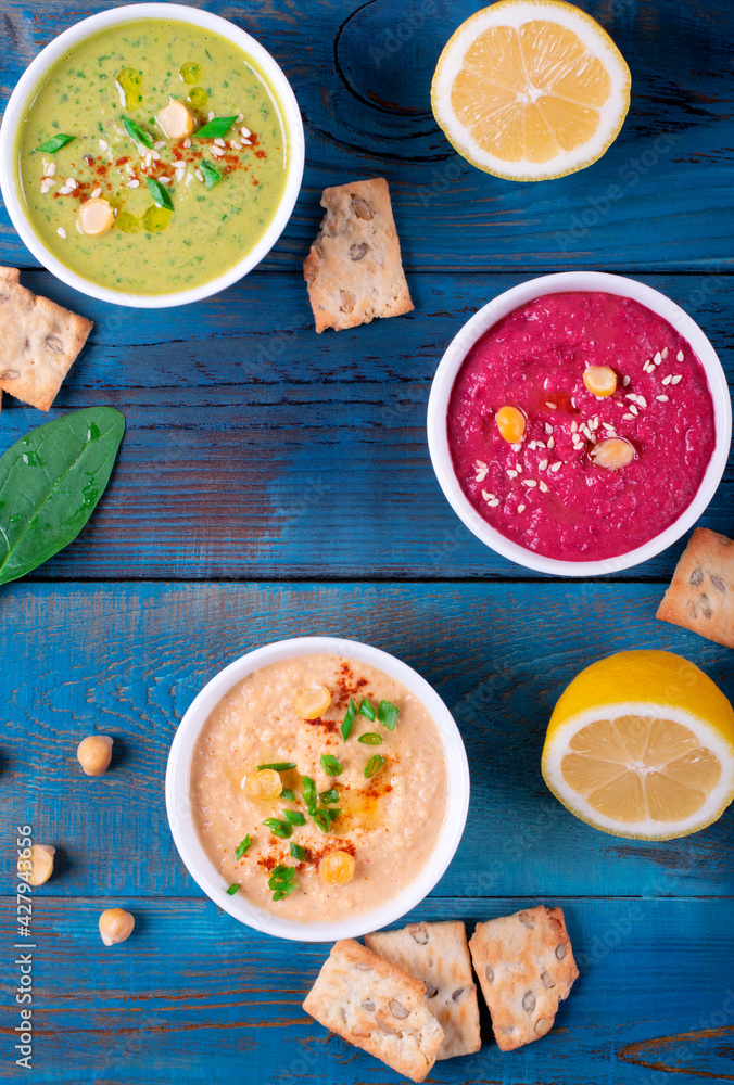 Colorful hummus in white bowls on blue wooden table. Classic, beetroot and spinach chickpea dips Colorful hummus in white bowls on blue wooden table. Classic, beetroot and spinach chickpea dips